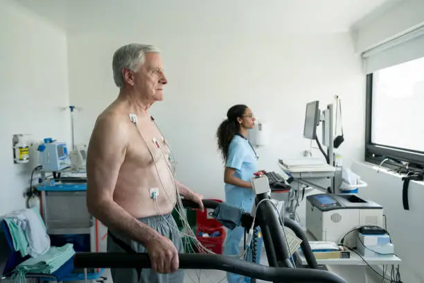 Photo of Senior man on a treadmill doing a stress test at the hospital while black nurse looks at the cardiac monitor