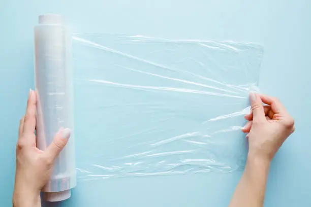 Photo of Woman's hand using a roll of transparent polyethylene food film for packing products on the pastel blue table. Empty place for text or logo.