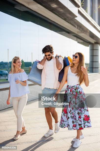 Junger Mann Alle Taschen Zu Tragen Während Zwei Freundinnen Sind Lachen Und Scherzen An Seiner Seite Stockfoto und mehr Bilder von Einzelhandel - Konsum