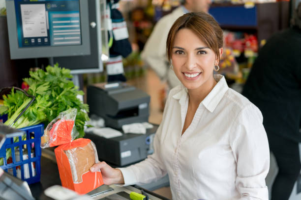 istock Friendly saleswoman at the cash register looking at camera smiling while scanning barcodes on products 955050126
