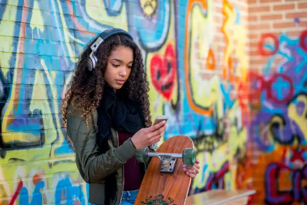 Photo of Teen girl with skateboard and mp3 stands in front of a graffiti brick wall