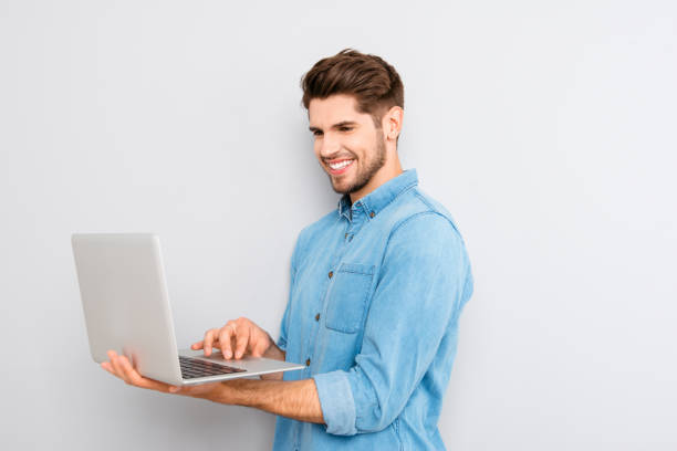 istock Smiling young man holding laptop and typing while standing against grey background 955084720