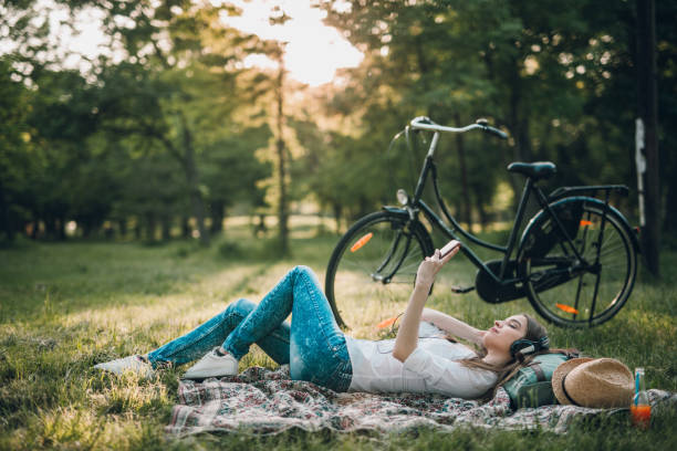 Cute young woman with headphones taking a rest from the bicycle ride, sitting on the blanket, listening to the music, enjoying springtime