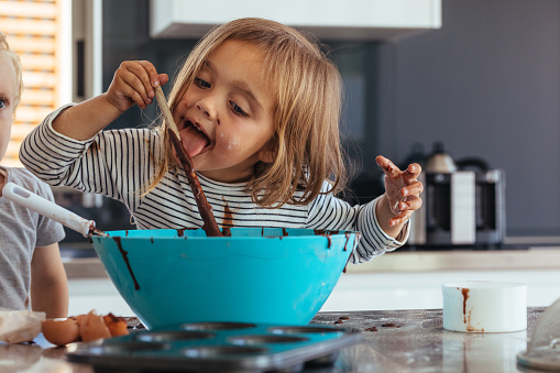 Little girl licking spoon while mixing batter for baking in kitchen  and her brother standing by. Cute little children making batter for baking.