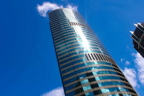 istock Modern sky scraper towering into blue sky with fluffy blue clouds and sun flare reflects surrounding builings in its curved facade 955092284