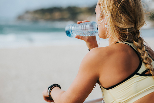 Over the shoulder photo of a blonde fitness girl drinking water and looking at the ocean