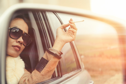 Confidence woman looking from side mirror of car and smoking cigarette with sunglasses in sunset