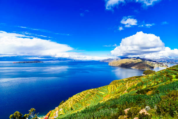 Terraced agricultural fields on Amantani Island, Peru, stretch across the hillside, overlooking the deep blue waters of Lake Titicaca under a vast sky with clouds