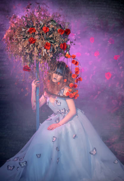 Cute young woman dressed in the blue ball gown. The woman with bizarre makeup and bouffant hairstyle is sitting next to the treelike floristic composition, she is embracing it. The woman's head in a bird's cage. Studio shooting on a background of a fir-tree wall with a rosebuds