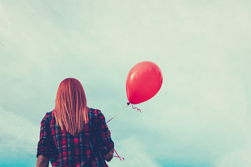Back view of red-haired girl holding red balloon against the sky.