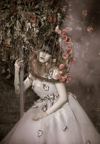 Sepia toned picture. The young woman dressed in the ball gown. The woman with bizarre makeup and bouffant hairstyle is sitting next to the treelike floristic composition, she is embracing it. The woman's head in a bird's cage. Studio shooting on a background of a fir-tree wall with a rosebuds
