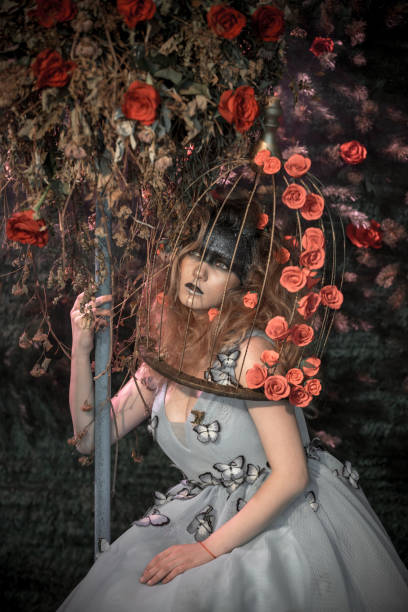The young woman dressed in the ball gown. The woman with bizarre makeup and bouffant hairstyle is sitting next to the treelike floristic composition, she is embracing it. The woman's head in a bird's cage. Studio shooting on a background of a fir-tree wall with a rosebuds