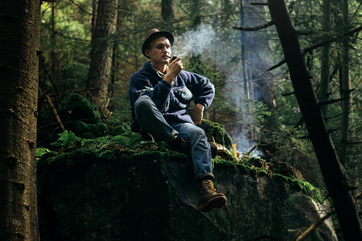 stylish hipster traveler smoking tobacco pipe in sunny forest in the mountains
