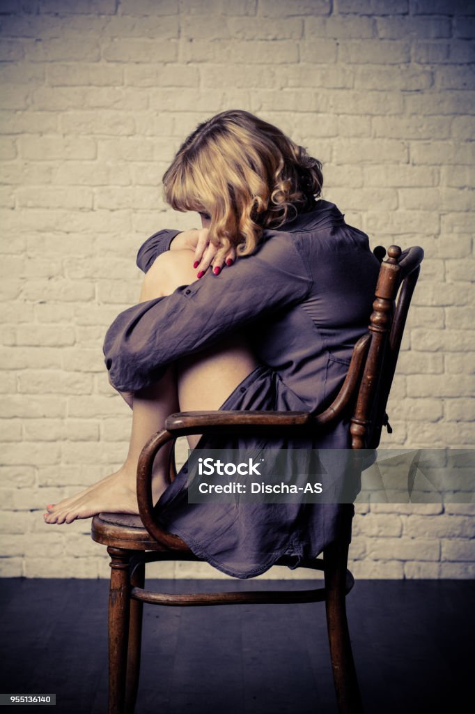 A young woman and a chair. Woman sitting on a wooden chair. His knees clasping his hands. The woman is wearing a long shirt. Accessibility Stock Photo