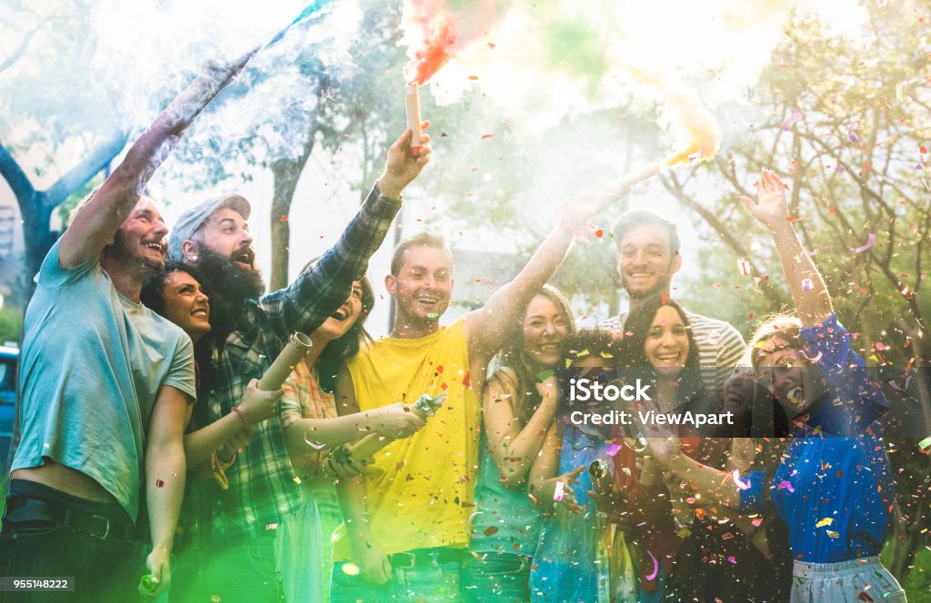 Happy friends having fun at garden party with multicolored smoke bombs outdoor - Young millennial students celebrating spring break together - Genuine youth concept - Focus on confetti and left people Party - Social Event Stock Photo