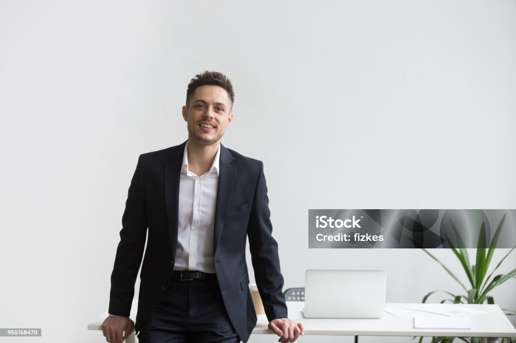 Smiling company CEO posing near office desk Smiling company CEO leaning back at office table posing near desk looking at camera satisfied with successful business. Concept of confidence, achievement, rewarding Men Stock Photo