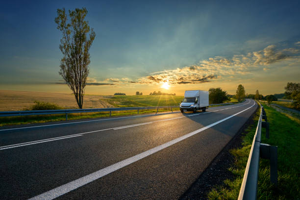 White delivery van driving on asphalt road around farm fields in rural landscape at sunset White delivery van driving on asphalt road around farm fields in rural landscape at sunset delivery van stock pictures, royalty-free photos & images