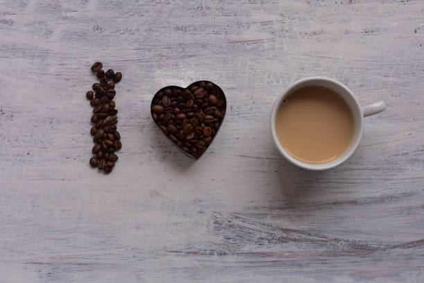 istock Cup op coffee and Heart shape roasted coffee beans on white rustic wooden table/ Romantic still life food design 955267240