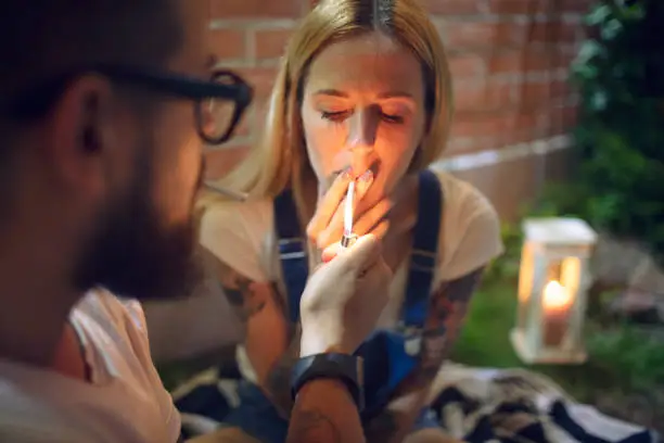 Bearded man lightning wife's cigarette with cigarette lighter.