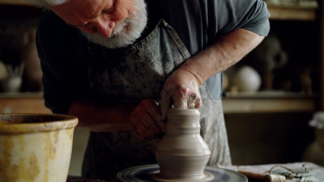 Hard-working senior potter is wetting hands in bowl with water and touching clay jar on spinning throwing-wheel. Bearded man is concentrated on work.