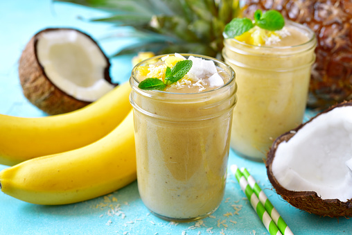 Tropical smoothies with banana, coconut and pineapple in a mason jars on a light blue slate, stone or concrete  background.