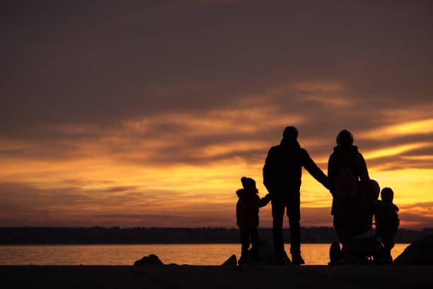 Young family with children holding hands silhouetted against an vivid orange ocean sunset.