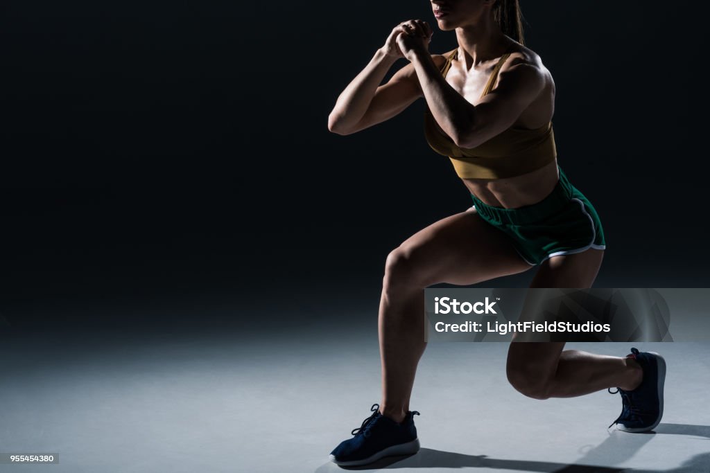 cropped view of sporty female bodybuilder doing lunges, on black Exercising Stock Photo