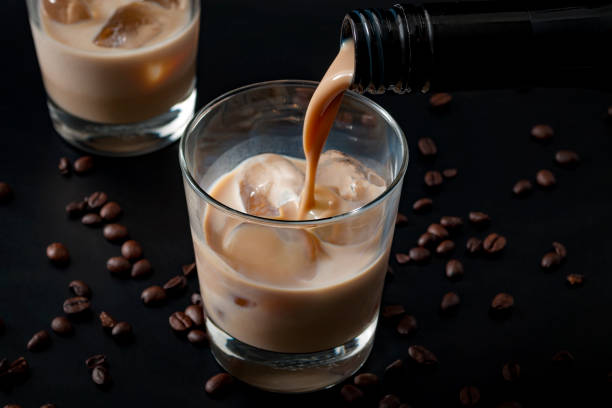 Coffee flavoured irish cream whiskey pouring in a glass Pouring irish cream in a glass with ice, surrounded by coffee beans on a dark black background cafe transparent glass table stock pictures, royalty-free photos & images
