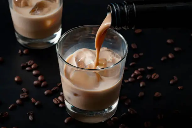 Pouring irish cream in a glass with ice, surrounded by coffee beans on a dark black background