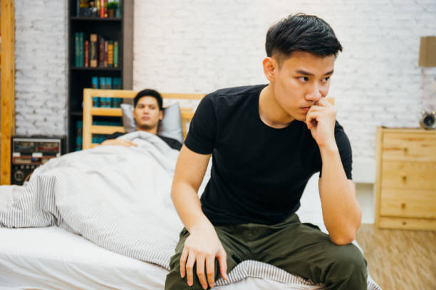 Asian gay couple having argument with each other in bedroom. Thoughtful gay man having stress while another is sleeping - Homosexual love concept Asian gay couple having argument with each other in bedroom. Thoughtful gay man having stress while another is sleeping - Homosexual love concept sad gay stock pictures, royalty-free photos & images