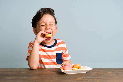 Cute child is eating macaroon on blue background. He is happy.