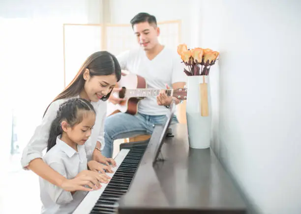 Photo of Asian family, Mother and daughter playing piano,Father playing guitar in family band at home, Concept for family relationship