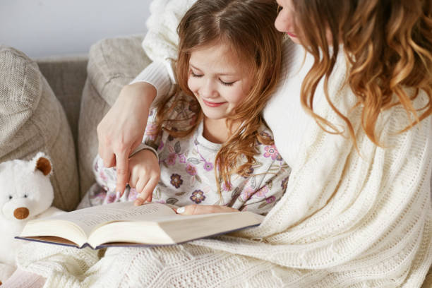 istock Happy Mother and daughter sitting on bed under plaid and reading book 955682884