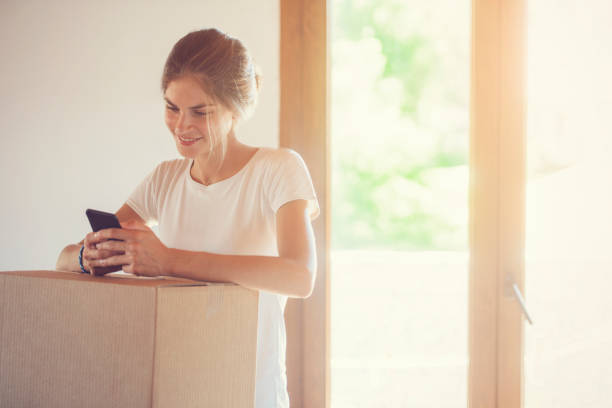 istock a beautiful young woman is communicating through her smart phone, next to a cardboard box. Calling for the mail delivery service. 955690464