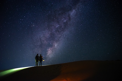 two travellers with flash light stand on sand dunes hill and look at the milky way on starry night sky , with copy space on sky