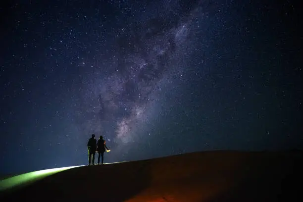 Photo of two travellers look at the milky way on starry night on sand dunes hill