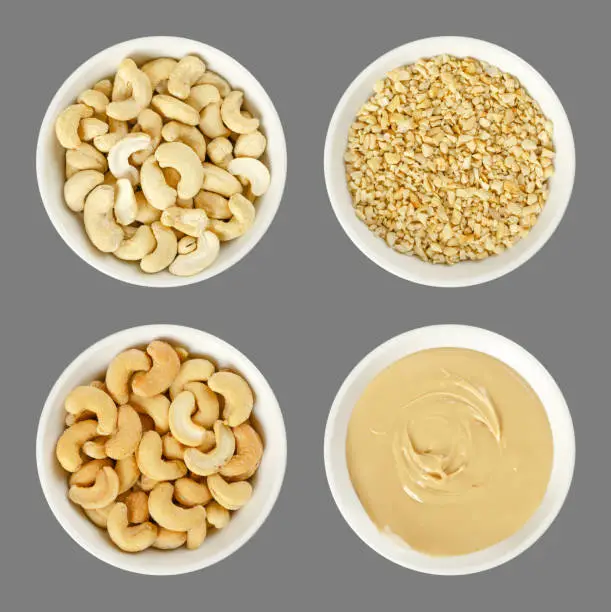 Cashew nuts in white porcelain bowls. Raw and processed nuts. Whole, roughly chopped, roasted, salted and as butter. Anacardium occidentale. Seeds. Isolated food photo, close up from above, over gray.