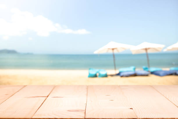 istock Wood table top on blur beach background with beach bag chairs and modern parasol 955772550