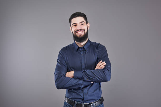 istock Young handsome man leaning against grey wall with arms crossed. A serious young man with a beard looks at the camera 955800992
