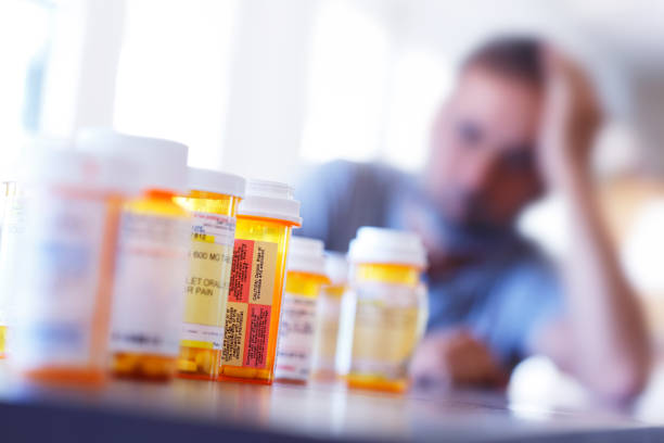 Medication Overload A large group of prescription medication bottles sit on a table in front of a distraught man who is leaning on his hand as he sits at his dining room table. The image is photographed with a very shallow depth of field with the focus being on the pill bottles in the foreground. junkie stock pictures, royalty-free photos & images