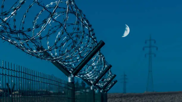 Photo of Close-up of sharp razor wire on fence. Gloomy blue sky and moon in background