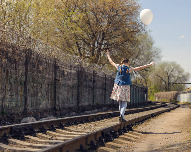 An attractive young woman holds a bunch of helium inflated balloons. She stands on railroad tracks in a run down, urban area. The wall behind her is full of graffiti.