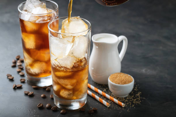 istock Ice coffee in a tall glass over and coffee beans on a old rustic wooden table. Cold summer drink on a dark background with copy space. The process of pouring drink from a coffee pot into a glass 955891400