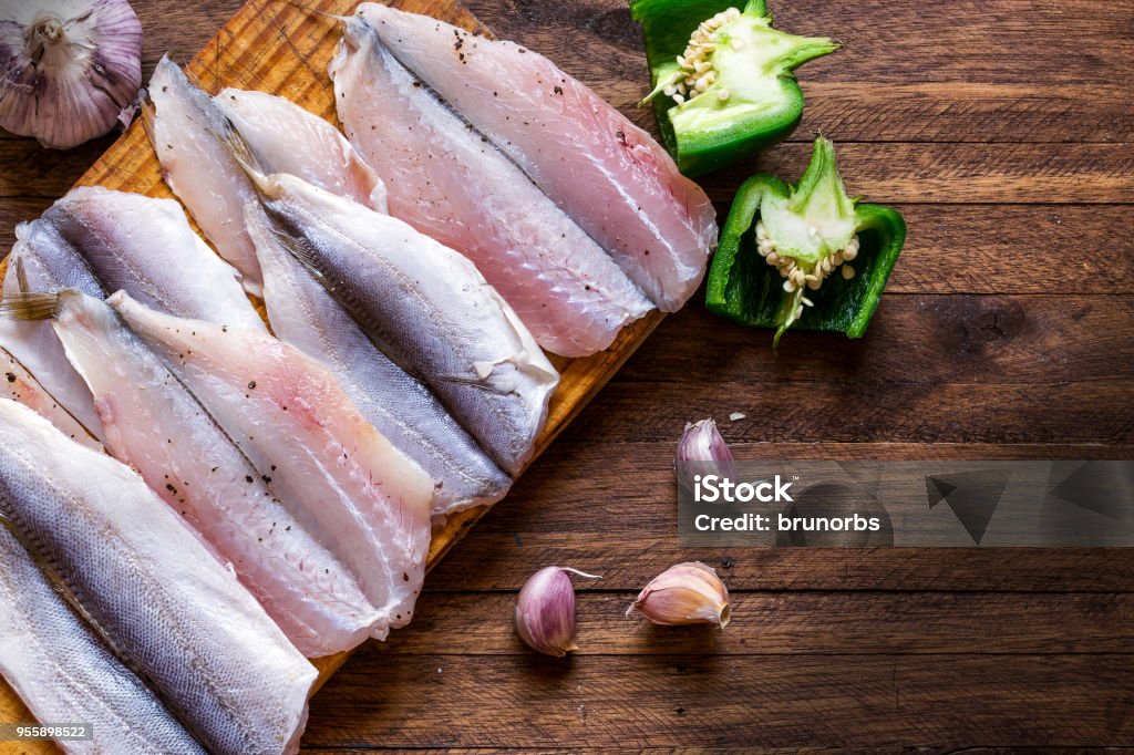 Délicieux et frais, épicés et assaisonnés les filets de poisson crus sur une planche à découper, sur un fond en bois, avec poivrons, entières et les gousses d’ail comme garniture - Photo de Poisson libre de droits