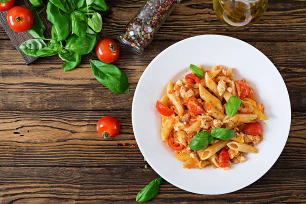 Penne pasta in tomato sauce with chicken, tomatoes, decorated with basil on a wooden table. Italian food. Pasta Bolognese. Top view