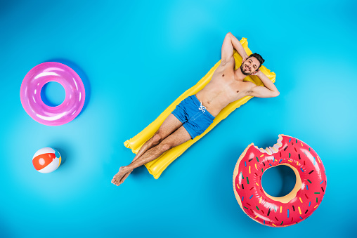 top view of handsome young man in shorts resting on inflatable mattress and smiling at camera on blue