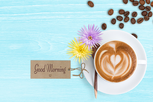 A cup of Latte coffee with good morning message, smiley, Gerbera flowers and scattered roasted coffee beans on pastel blue desk. Good morning coffee.