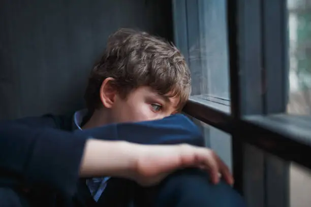 Photo of Pensive sad boy teenager in a blue shirt and jeans sitting at the window and closes his face with his hands.