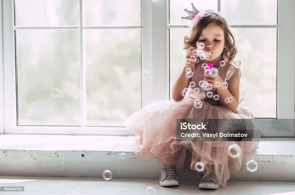 Little cute girl in dress Little cute girl in beautiful dress is sitting near the window at home and blowing soap bubbles. Child Stock Photo