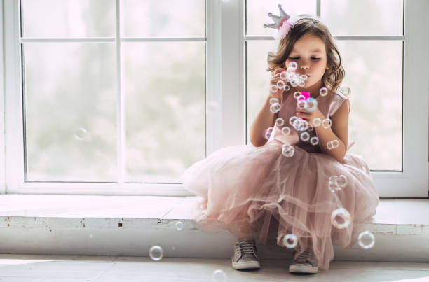 Little cute girl in dress Little cute girl in beautiful dress is sitting near the window at home and blowing soap bubbles. headwear photos stock pictures, royalty-free photos & images